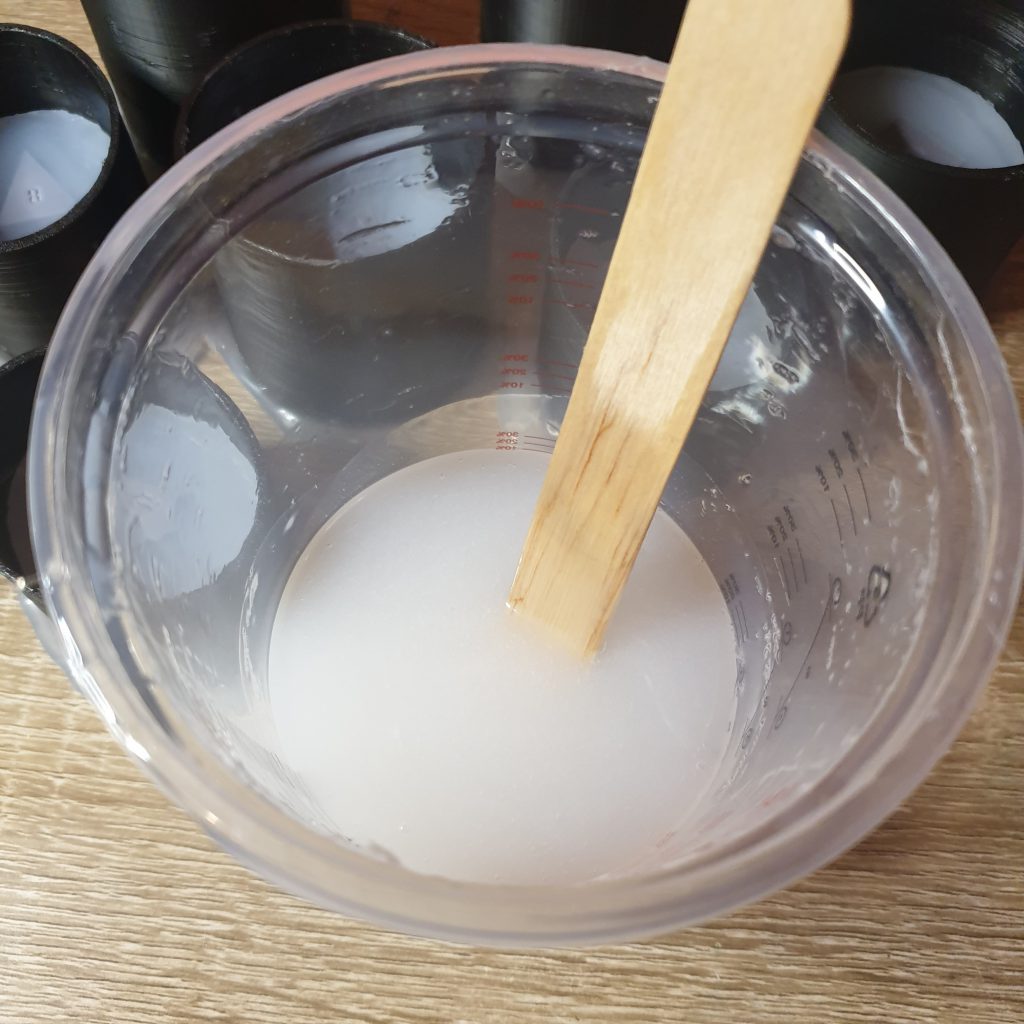 Silicone in a mixing cup with a large flat popsicle/lolly stick in with it. The dice moulds the silicone is going in are in the background.